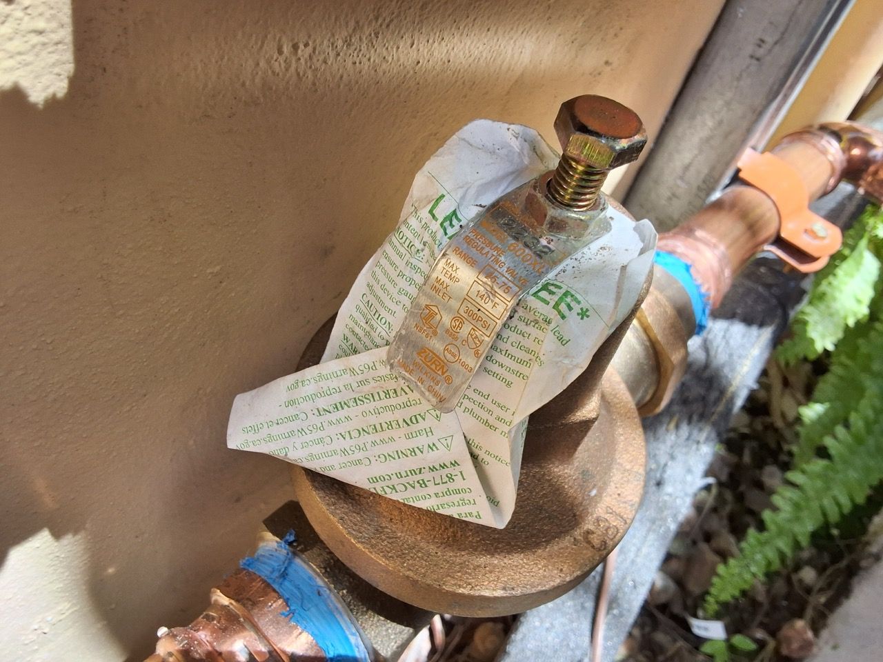 Tag hanging on an outdoor pipe valve beside a wall.