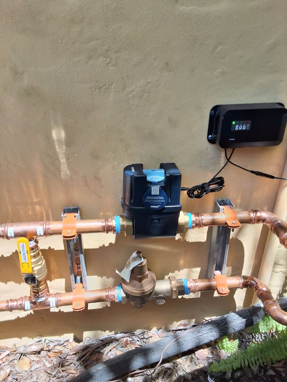 Outdoor water piping with valves, a pump, and a control box against a wall.