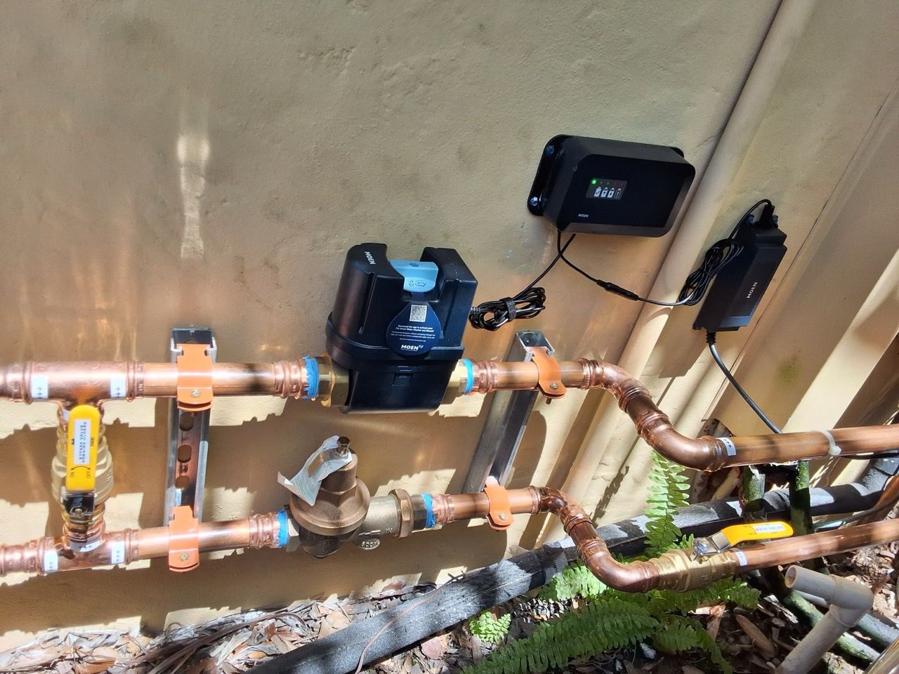 Outdoor plumbing pipes with a black water pump, valves, and electrical controls mounted on a wall.