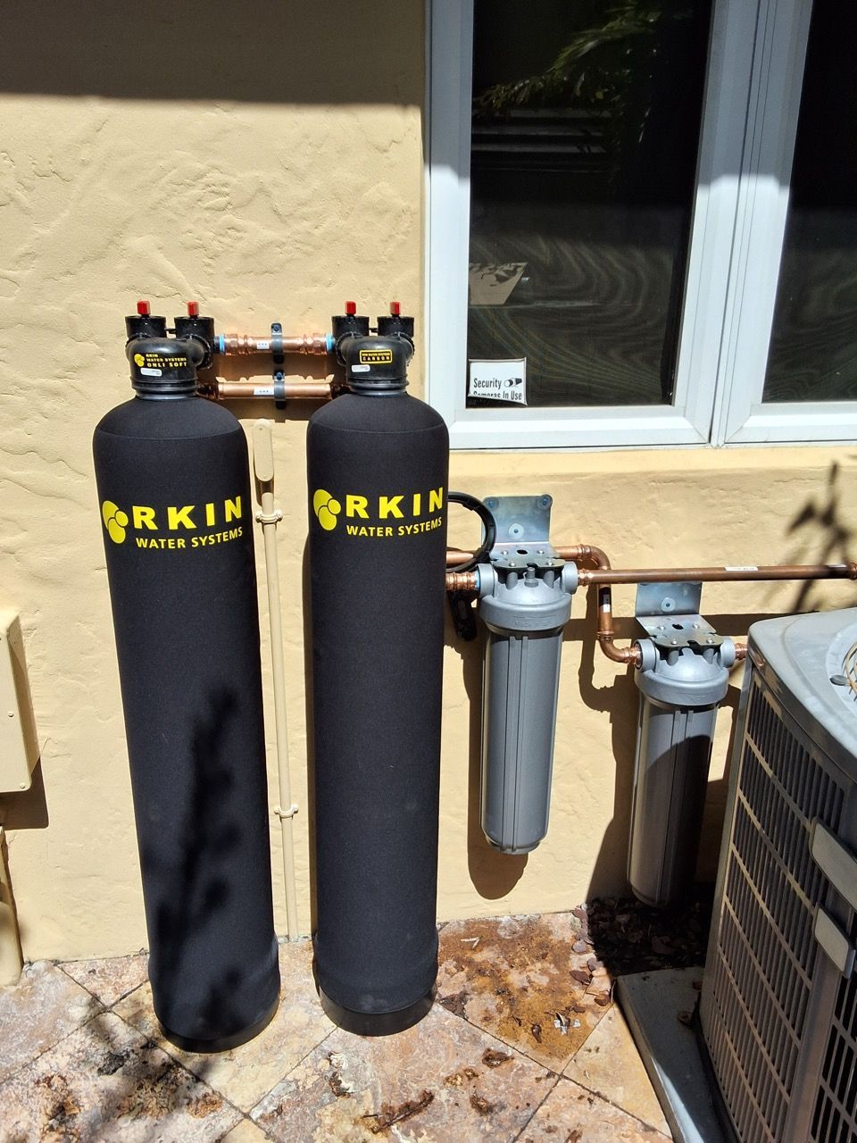 Two black cylindrical filters beside a house wall and window, with utility pipes and outdoor HVAC units nearby