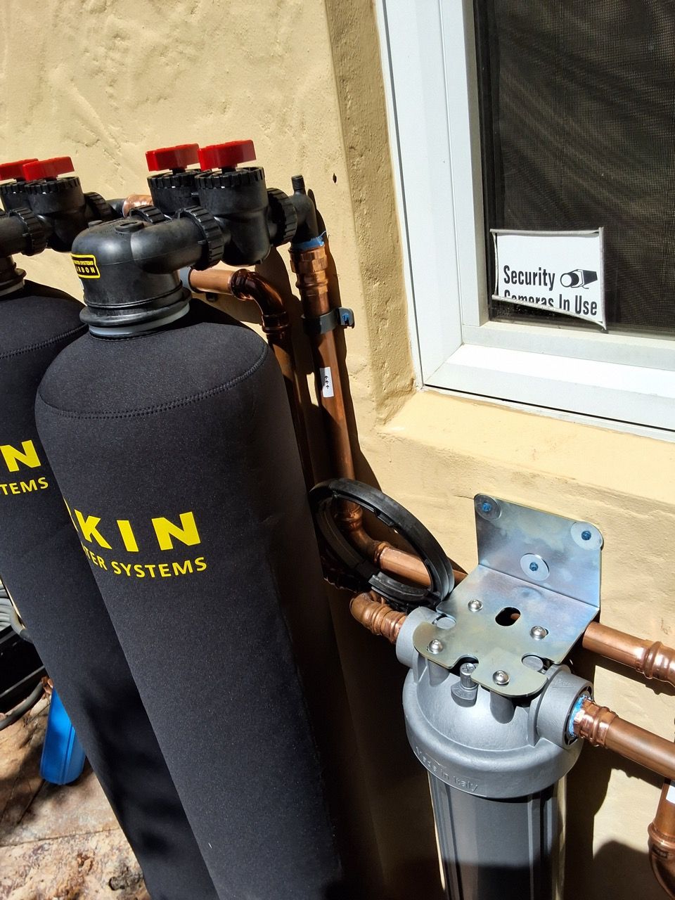 Black water softener tank with copper pipes and valves beside a window and wall