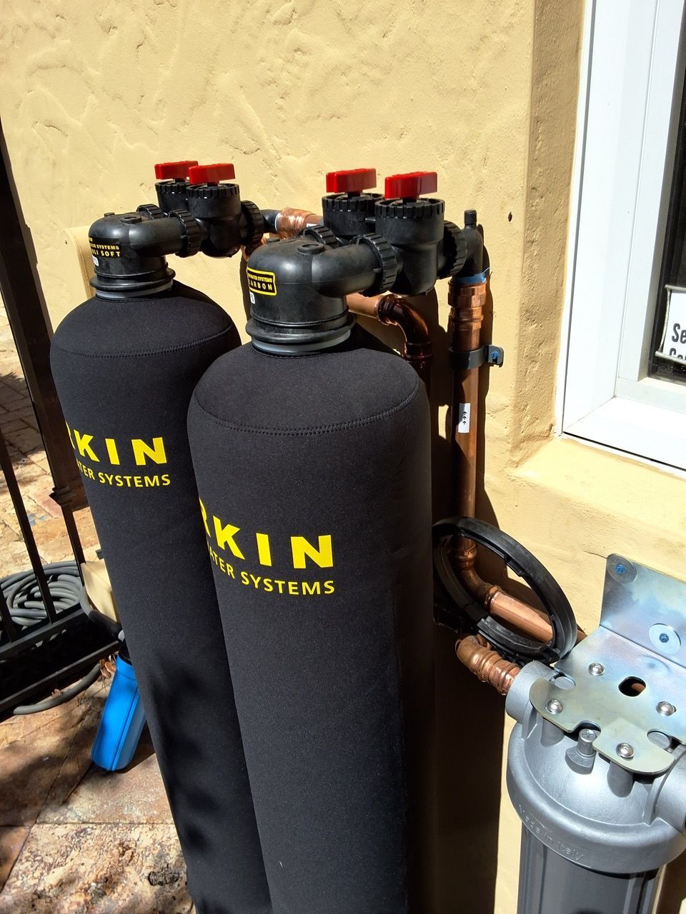 Two black water softener tanks with red valves and piping beside a beige exterior wall.