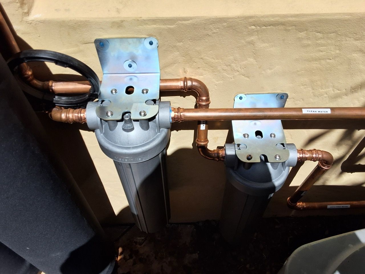 Copper pipes and two gray water filters mounted on a beige wall with black tubing nearby