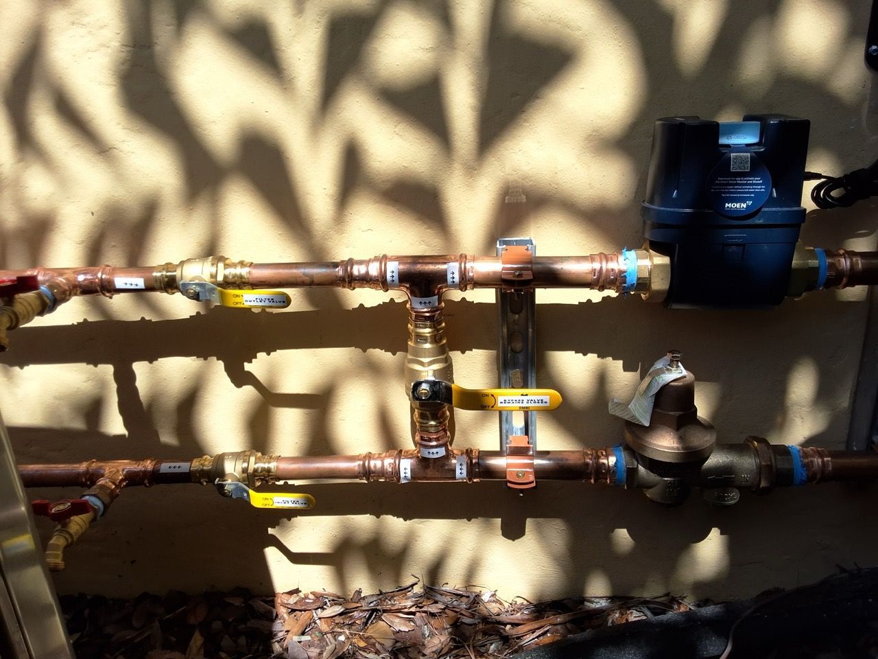 Outdoor copper water pipes with valves and a blue meter against a wall and ground shadow patterns