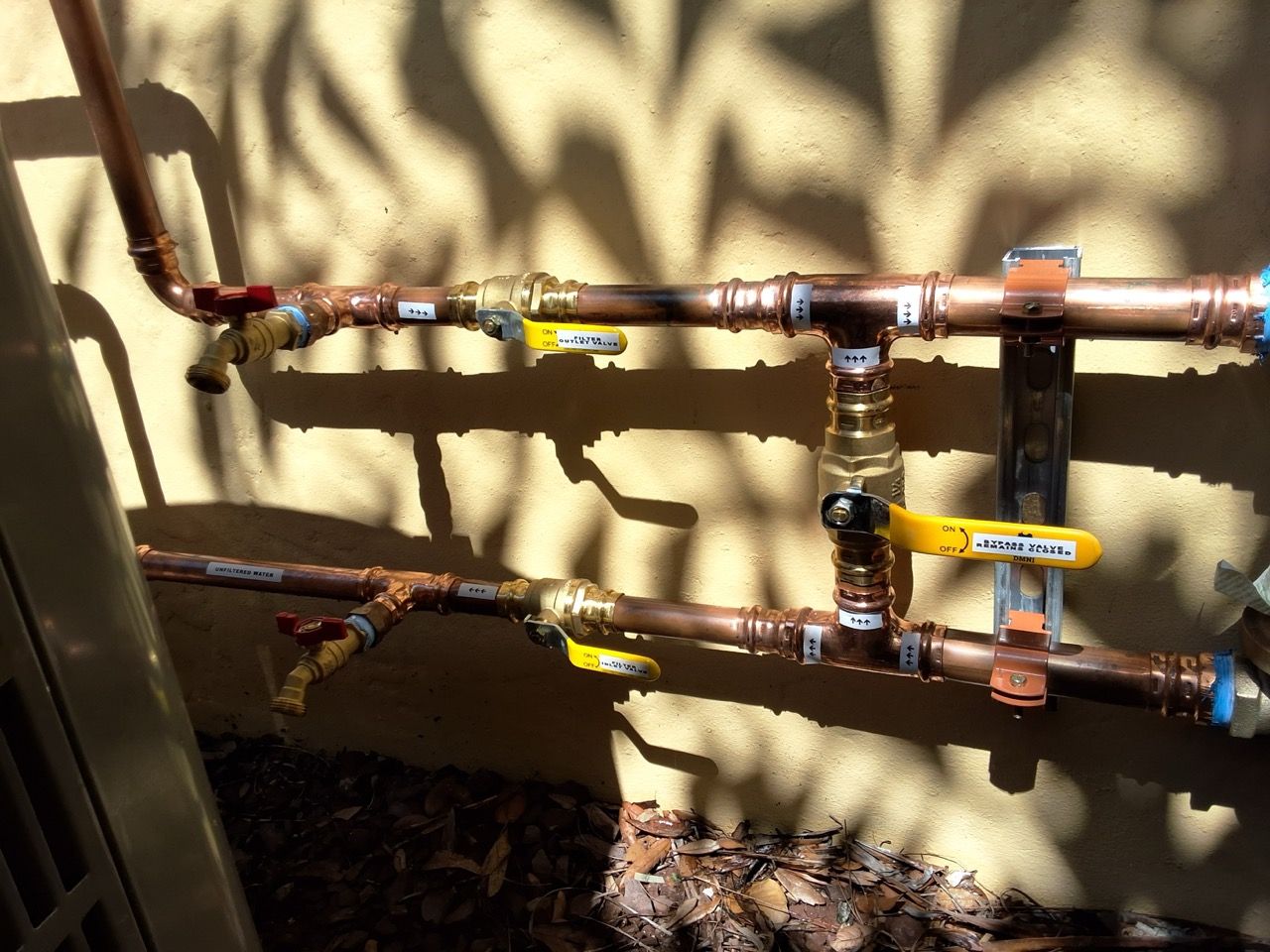 Outdoor copper water pipes with yellow shutoff valves mounted on a stucco wall in sunlight