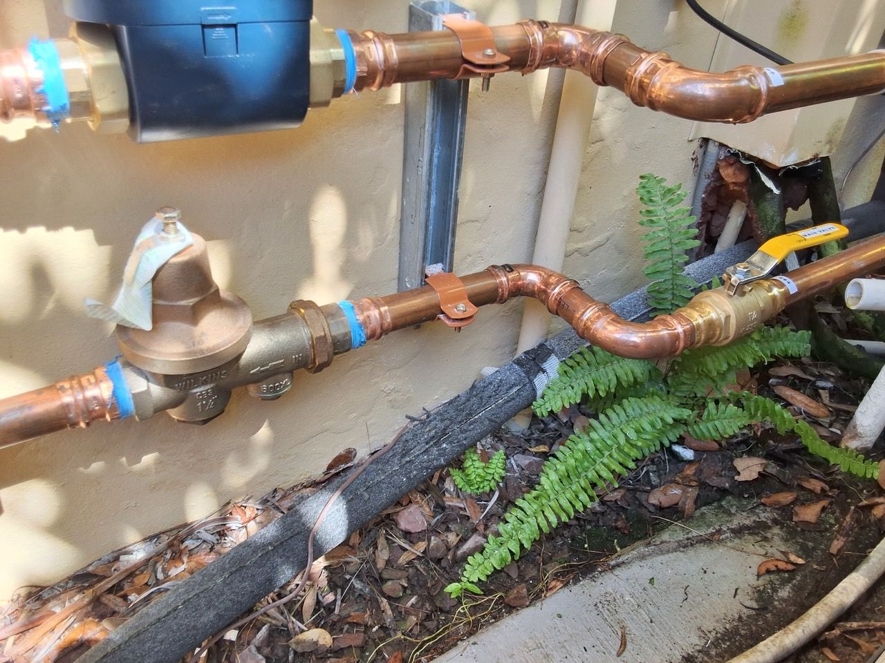 Copper pipes and valves along an exterior wall, with a black meter and plants below.