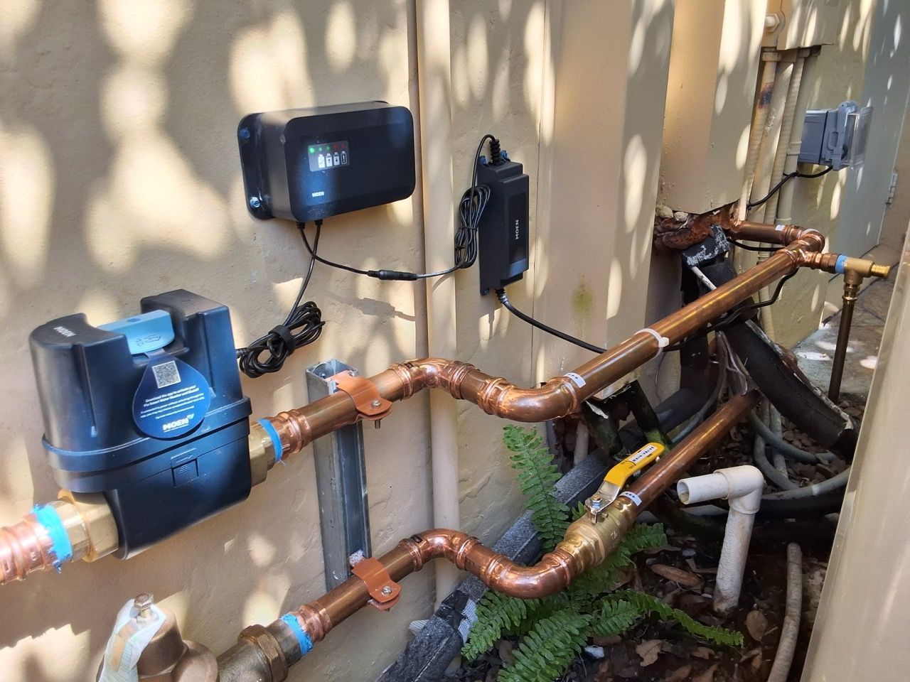 Outdoor plumbing pipes and utility meters mounted on a beige wall with copper piping and greenery below