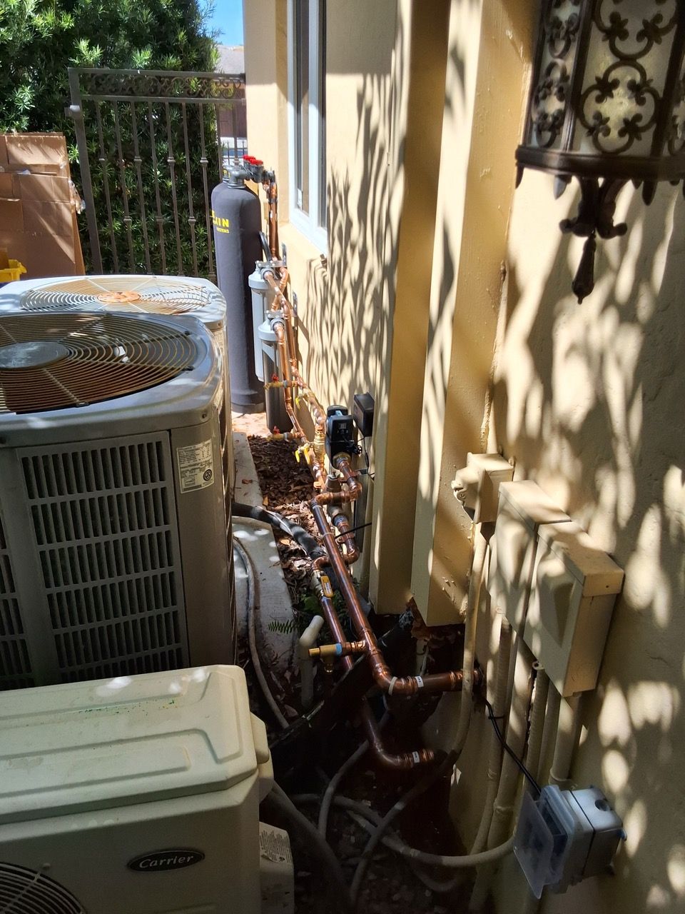 Outdoor HVAC units and utility pipes along a beige house wall in a narrow side yard.