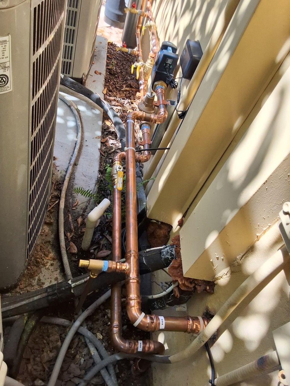 Tight outdoor utility area with copper pipes, hoses, and valves beside a building wall and fence.