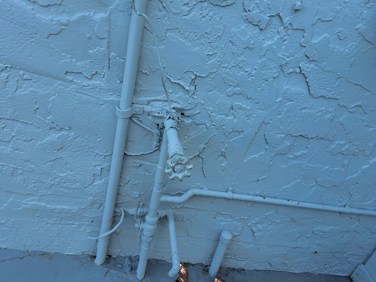 White stucco wall with exposed white pipes and cracked plaster near the ground