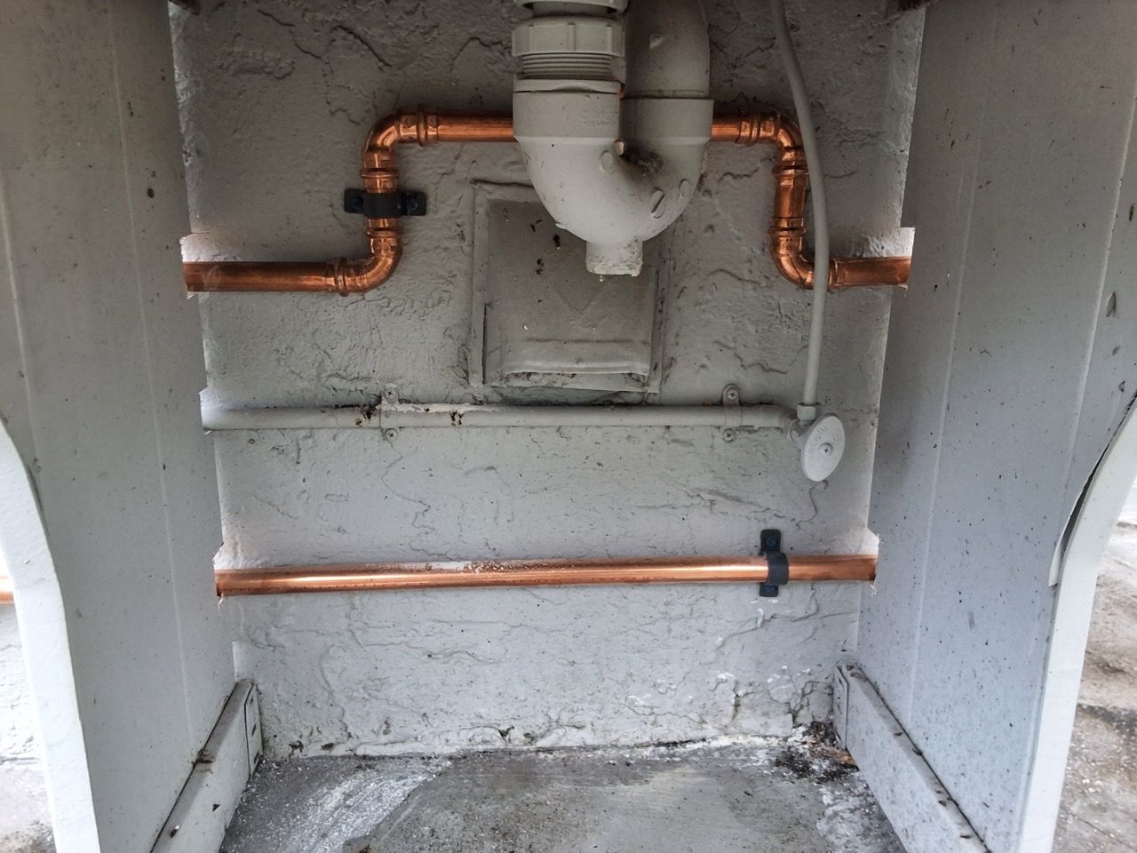 Copper pipes and a sink drain against a rough concrete wall in a small utility space