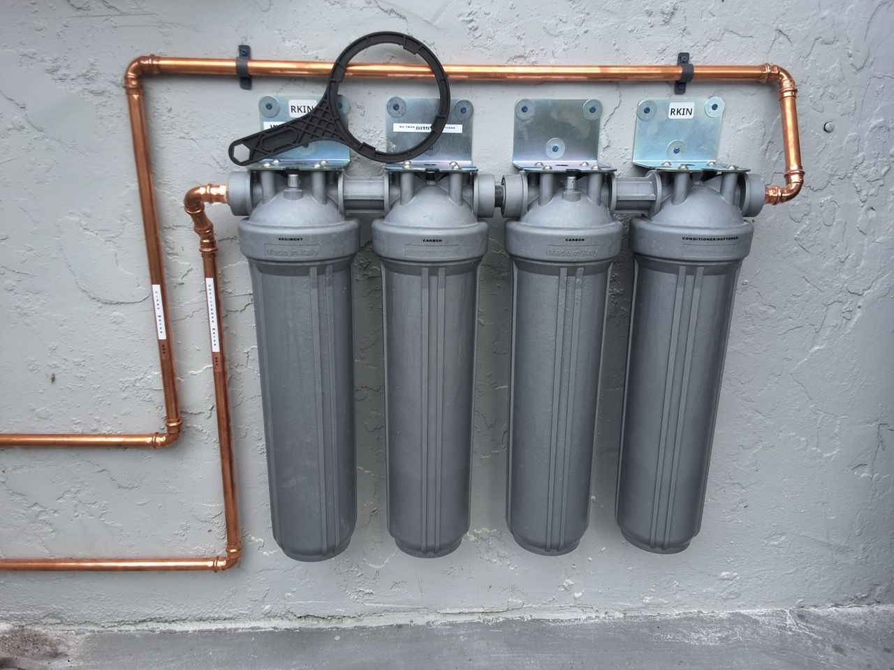 Four gray water filter housings mounted on a wall with copper pipes and a pressure gauge