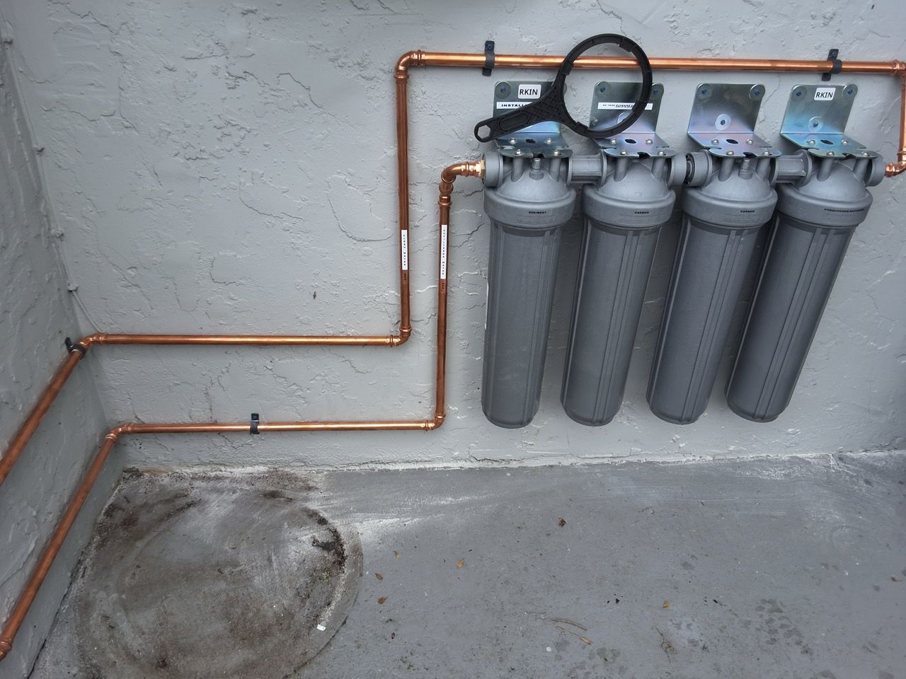 Wall-mounted water filter system with four gray canisters and copper piping in a concrete utility area