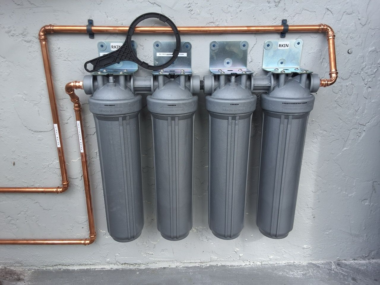 Four gray water filter canisters mounted on a wall with copper piping above and beside them
