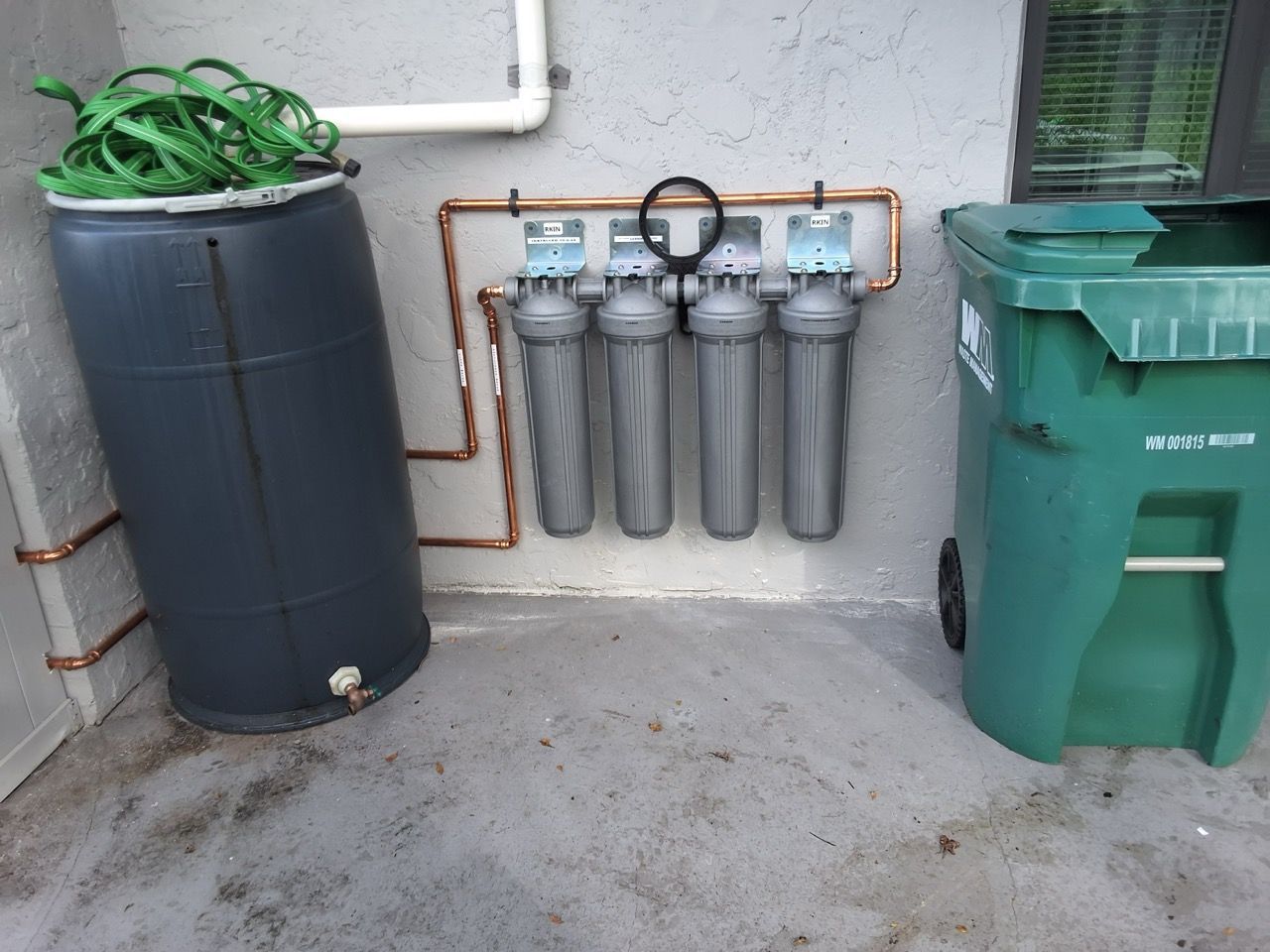 Water softener tanks and filters beside a wall, with a green trash bin and black rain barrel in a utility area.