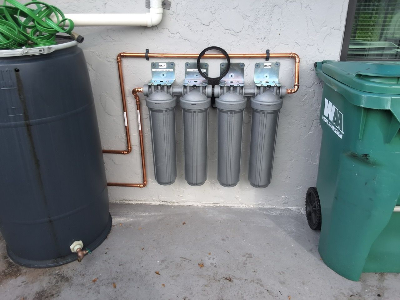 Water filtration system with three gray canisters mounted on a wall beside large trash bins.