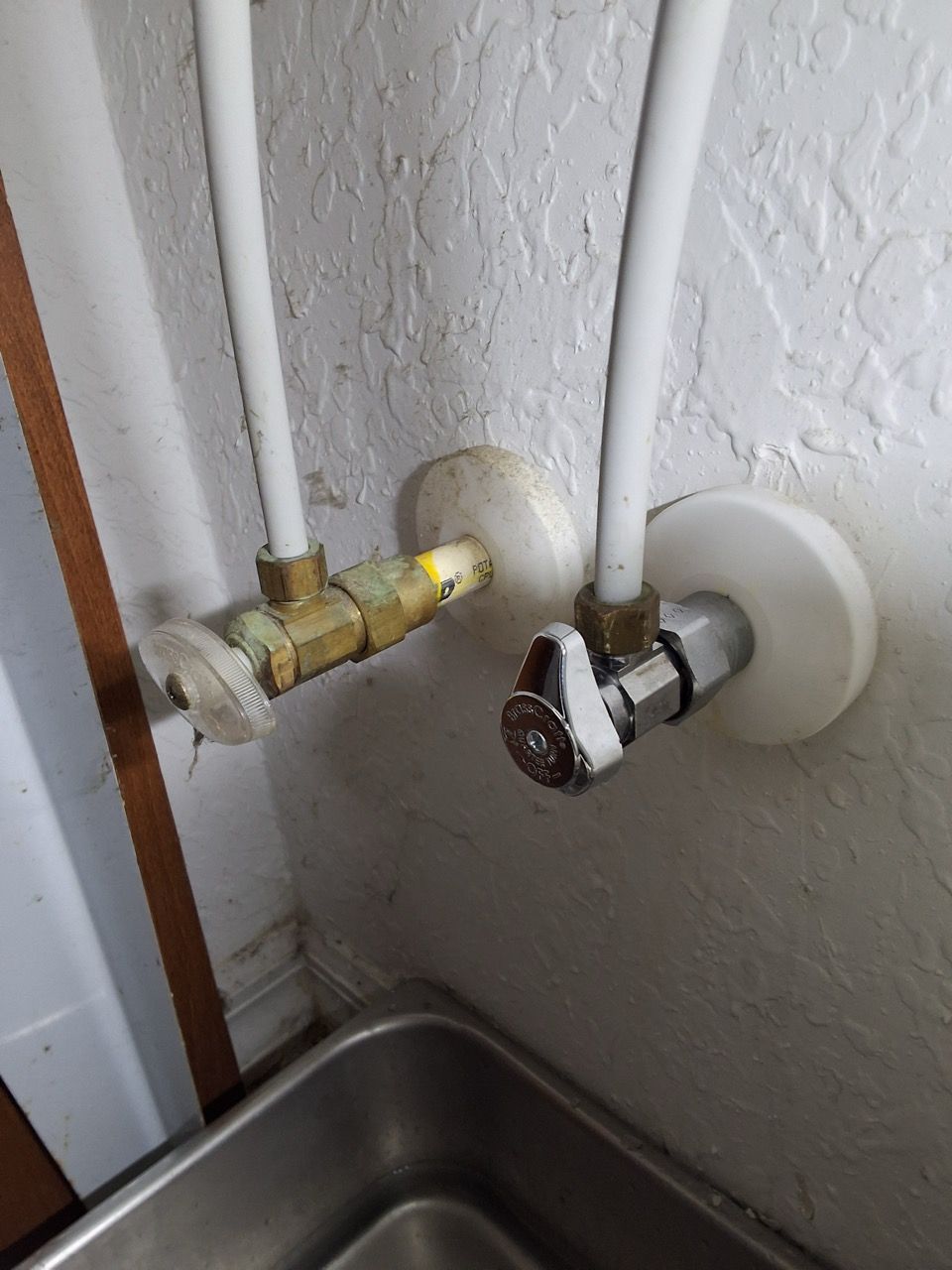 Close-up of plumbing valves and white pipes above a stainless steel sink against a textured wall.