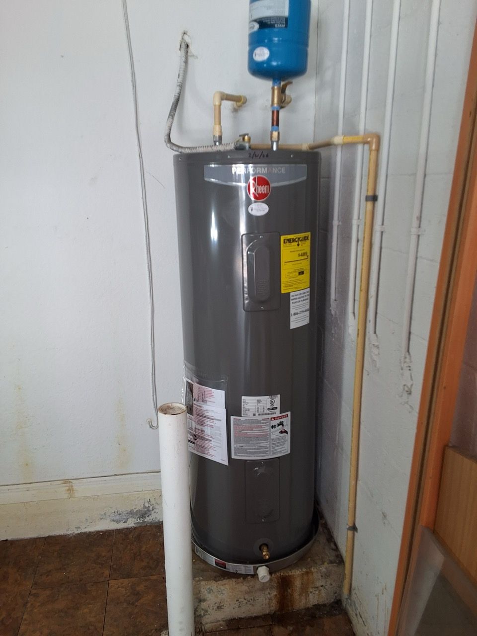 Gray water heater in a utility closet with pipes and a blue expansion tank above it.