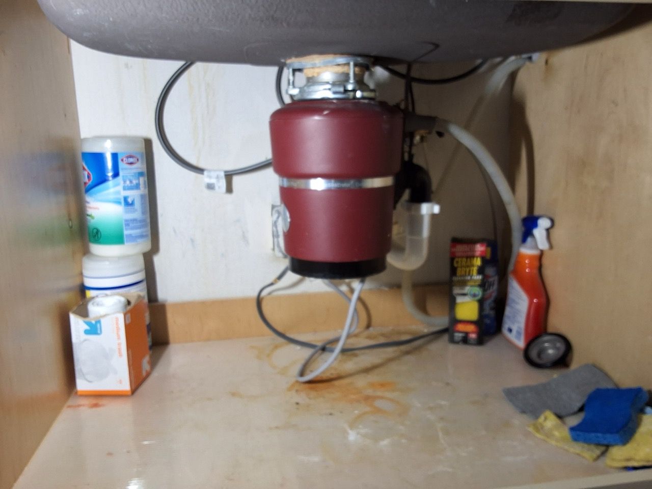 Under-sink cabinet with a red garbage disposal, cleaning bottles, and supplies on the floor
