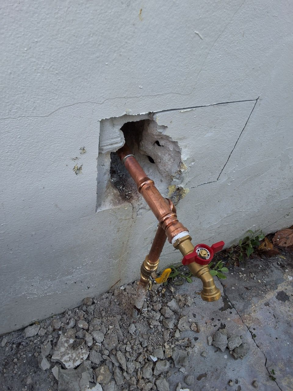 Copper pipes and a red valve exposed through a hole in a wall, with gravel and dirt below.