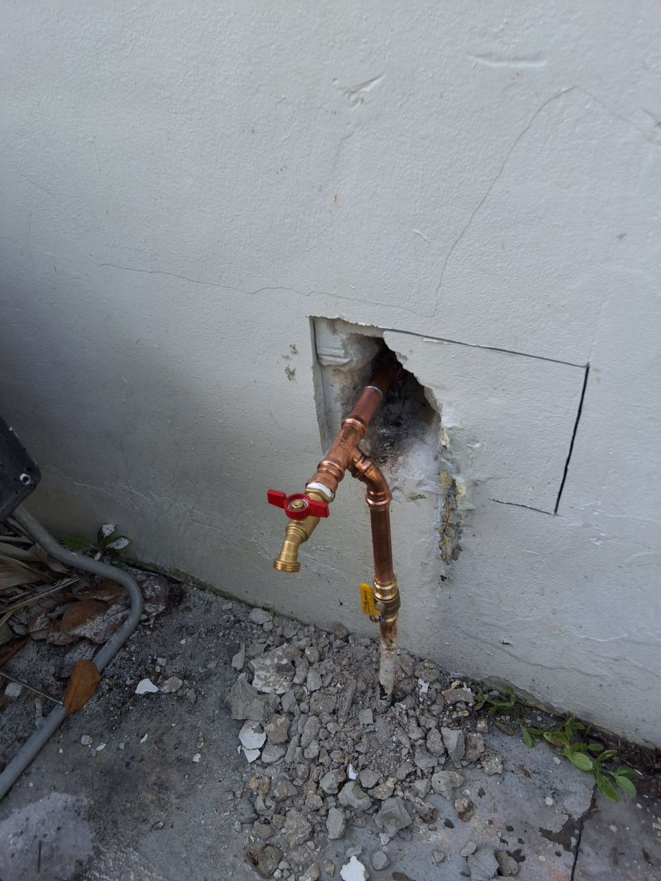 Damaged exterior wall with exposed copper pipes and two red-and-yellow shutoff valves near the ground