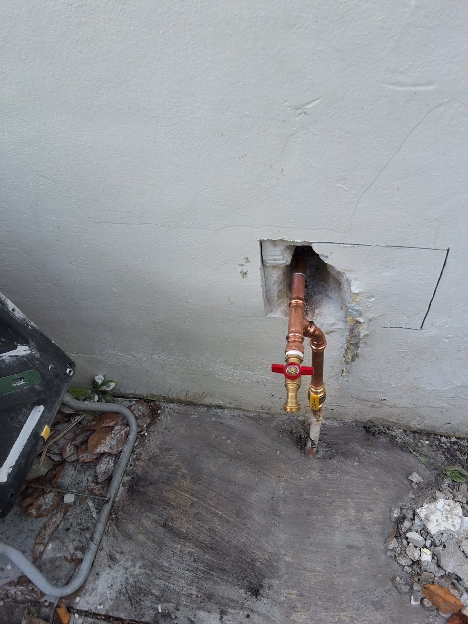 Outdoor wall with exposed copper pipes and red/yellow shutoff valves beside a concrete utility area.