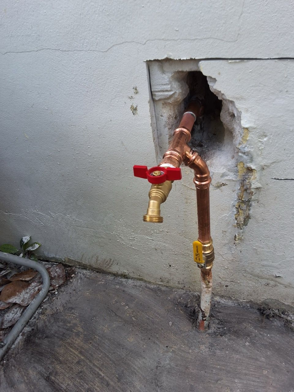 Copper pipes with a red shutoff valve protrude from a hole in a concrete wall.