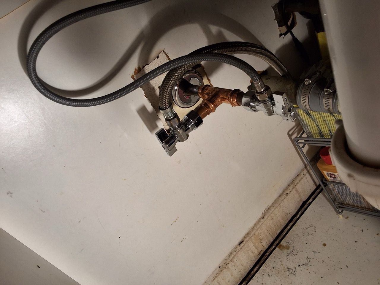 Close-up of tangled cables and metal plumbing parts against a white wall and floor corner.