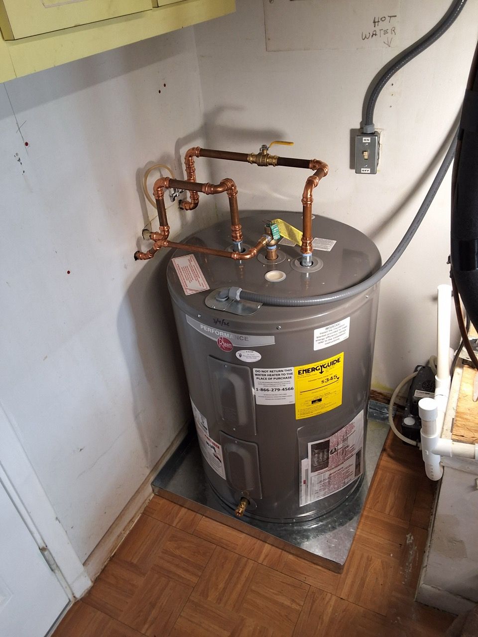 Water heater in a small utility closet with copper pipes and a power cord on the wall.
