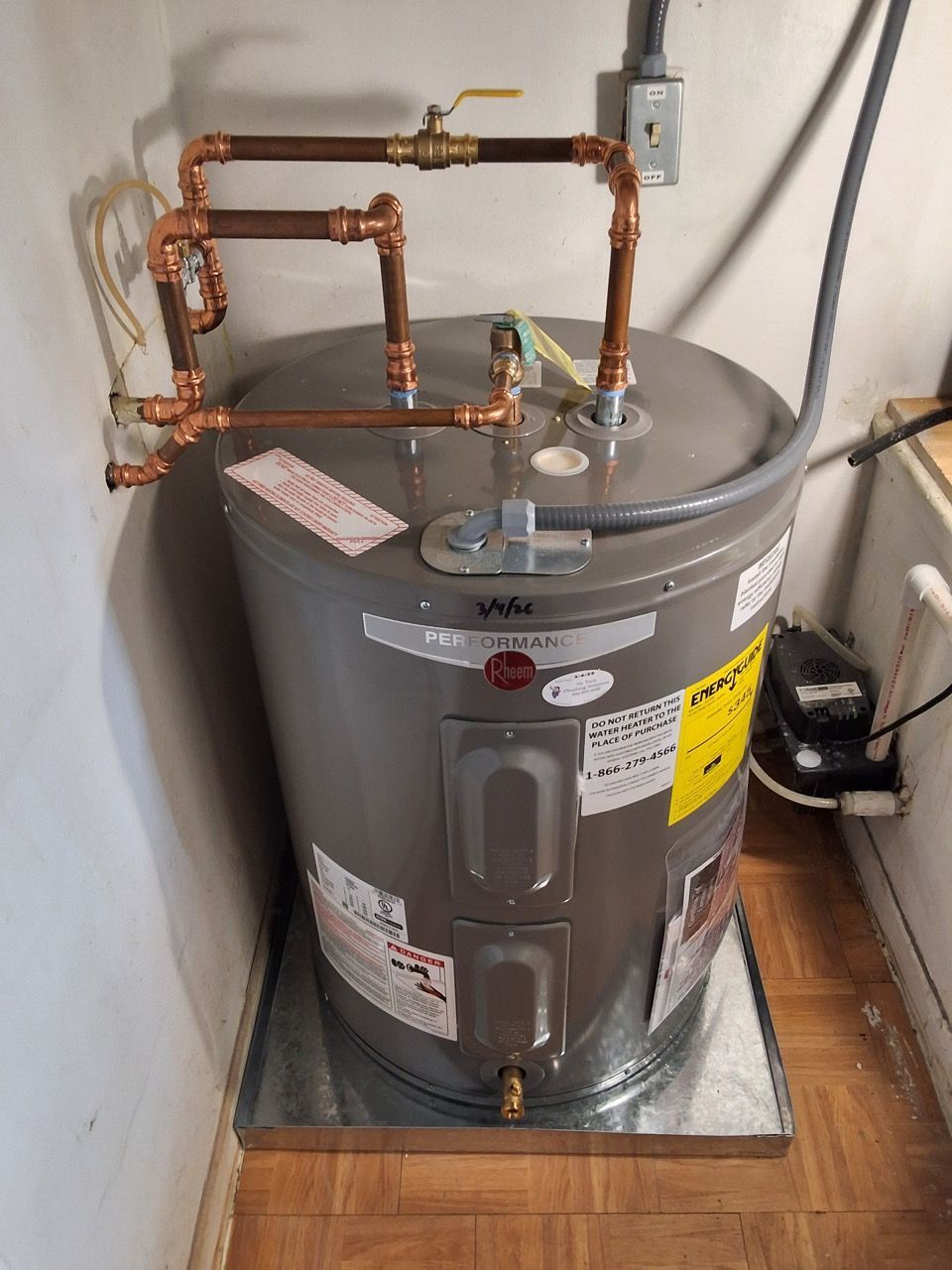 Water heater tank with copper pipes and valves in a small utility closet, sitting on a metal base.