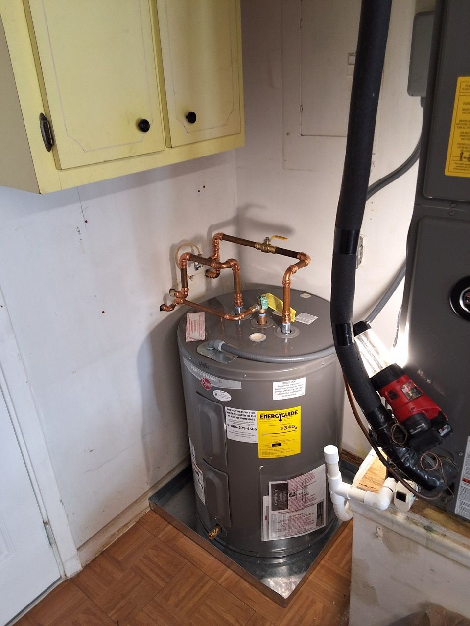Water heater with copper pipes in a small utility closet, next to a yellow cabinet and black furnace.