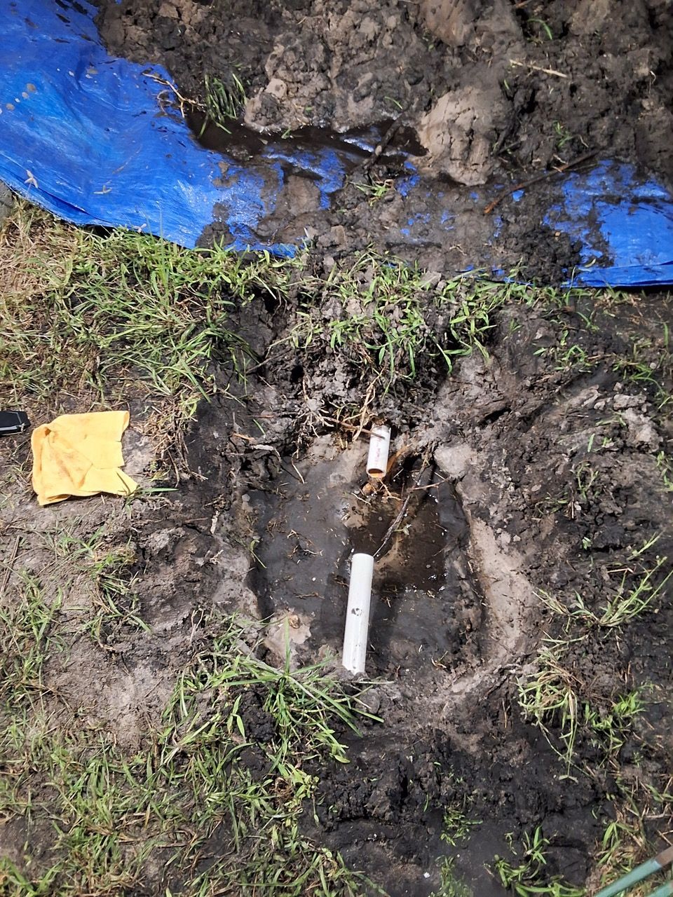 White pipe protruding from muddy ground beside blue tarp and small yellow cloth.