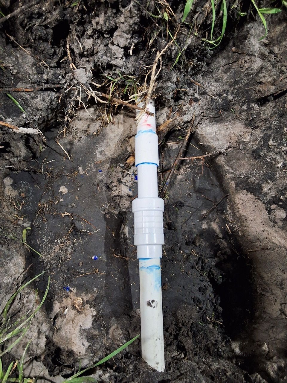 White plastic pipe with blue bands inserted into muddy ground near a small hole.