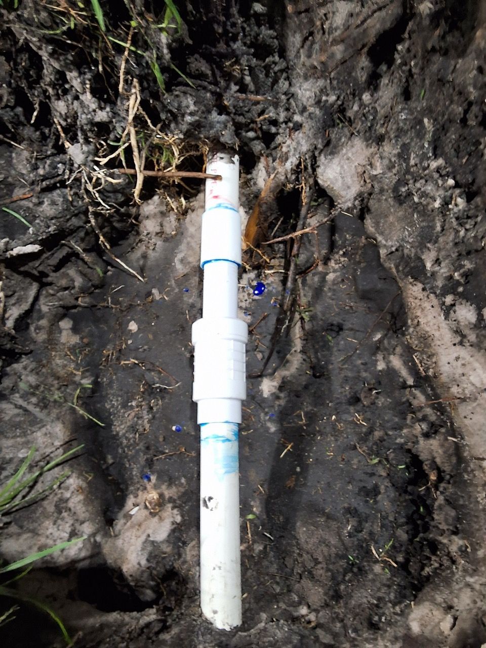 White PVC pipe stands upright against a dirt embankment with exposed roots.