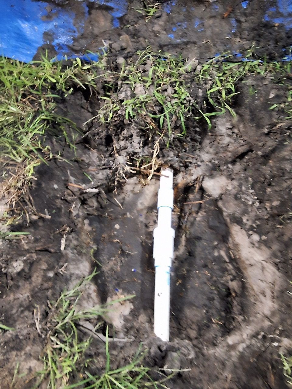 White PVC pipe in a dirt trench beside grass and blue plastic sheeting.