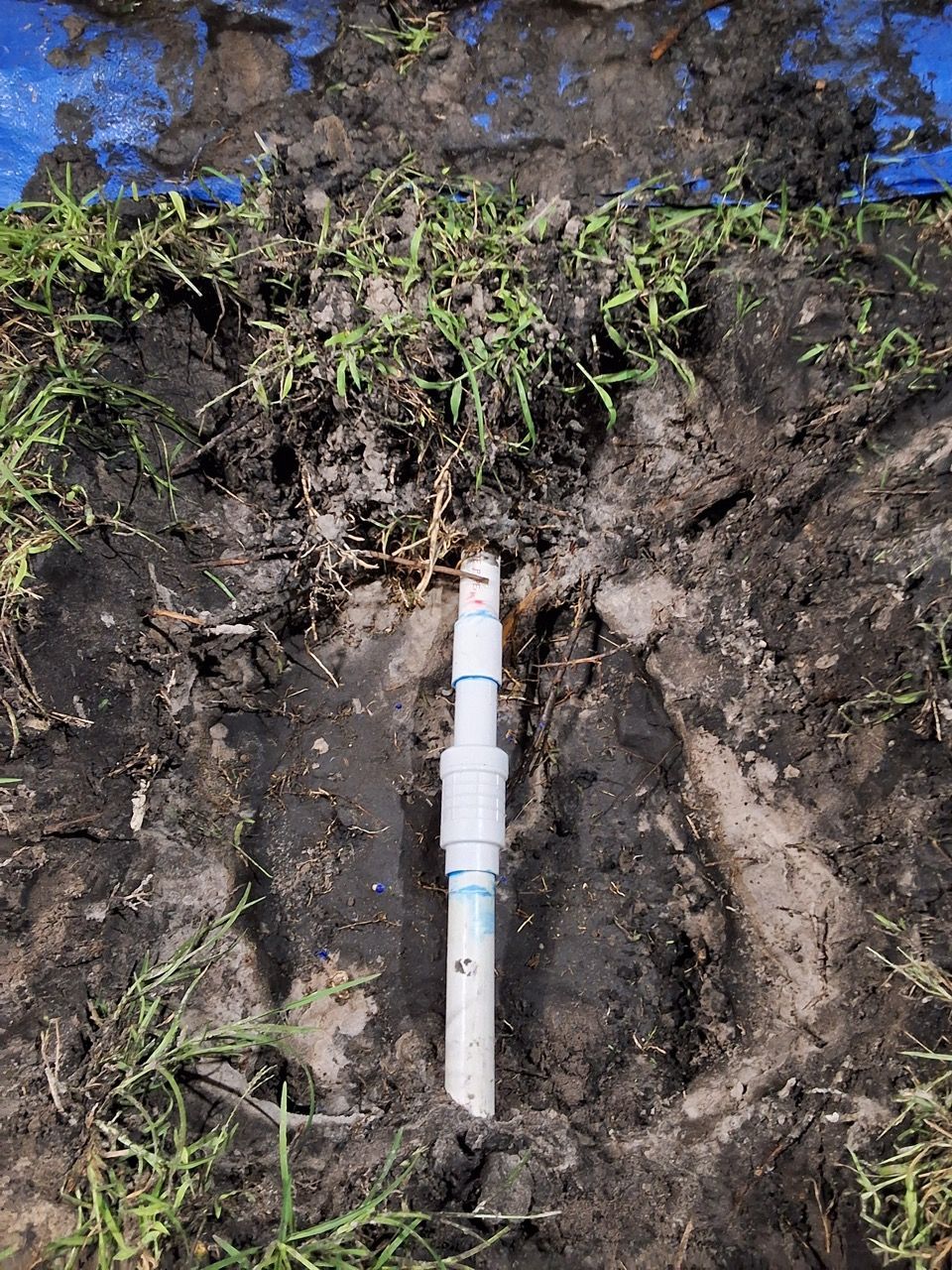 A white pipe fitting in a dirt trench, with exposed roots and blue tape along the soil edge.