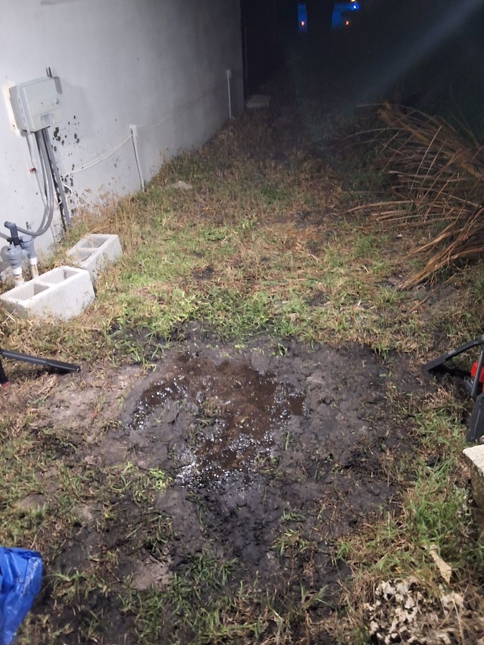 Narrow muddy backyard path beside a wall, with patchy grass and scattered debris at night.