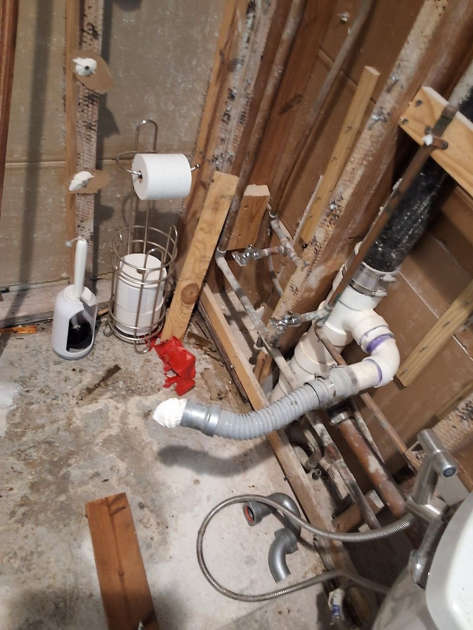 Exposed plumbing and sewer pipes in an unfinished wall with insulation and concrete floor debris