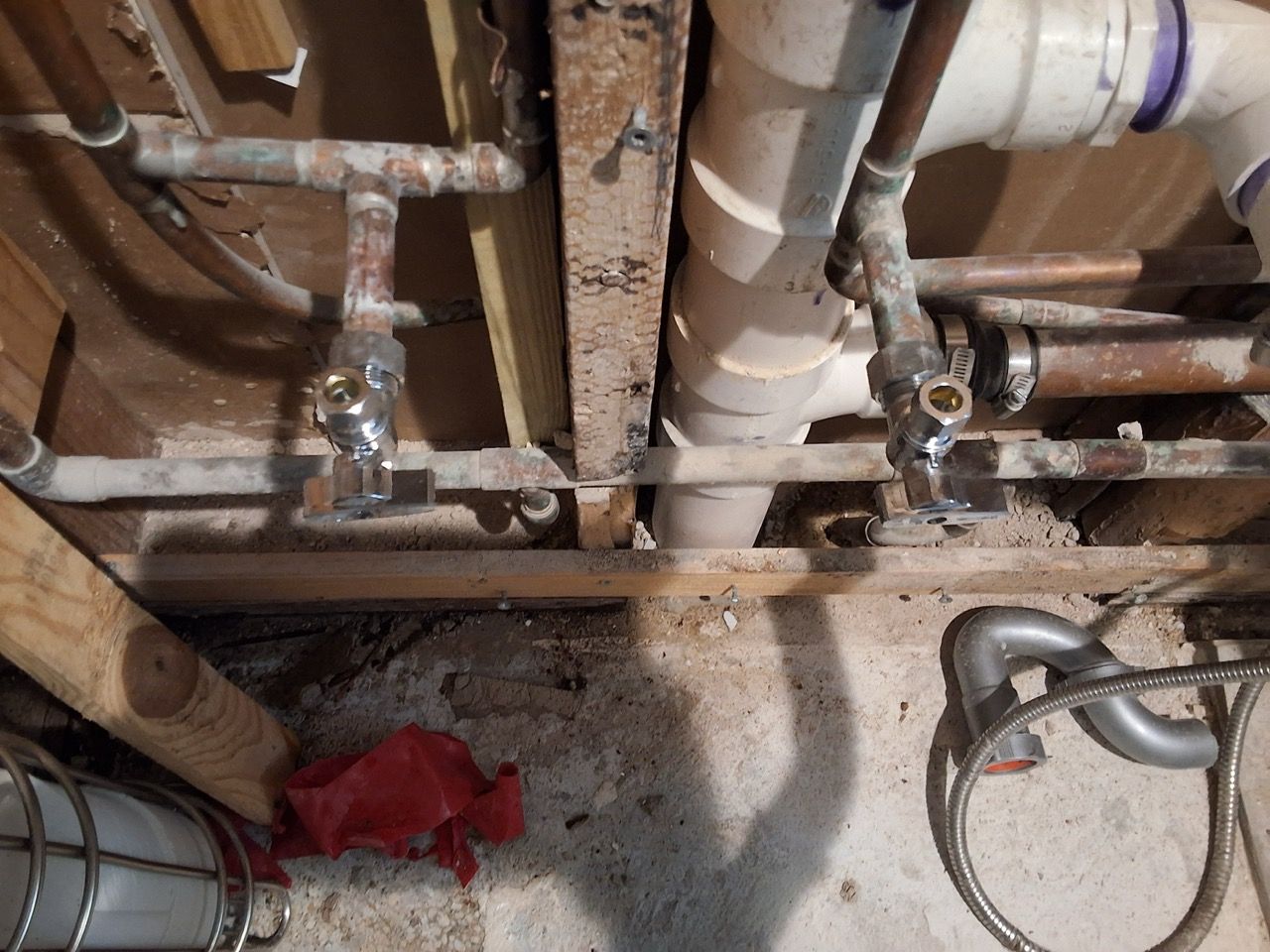 Under-sink plumbing with exposed pipes, valves, and a red cloth on the floor.