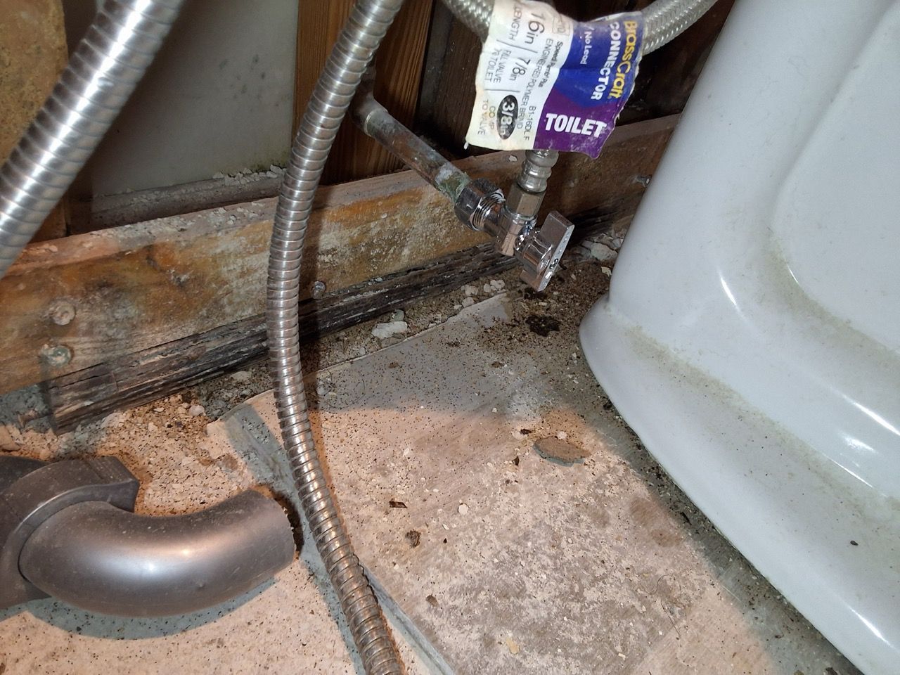 Under-sink plumbing with a flexible hose, PVC pipe, and stained floor pan near a toilet base