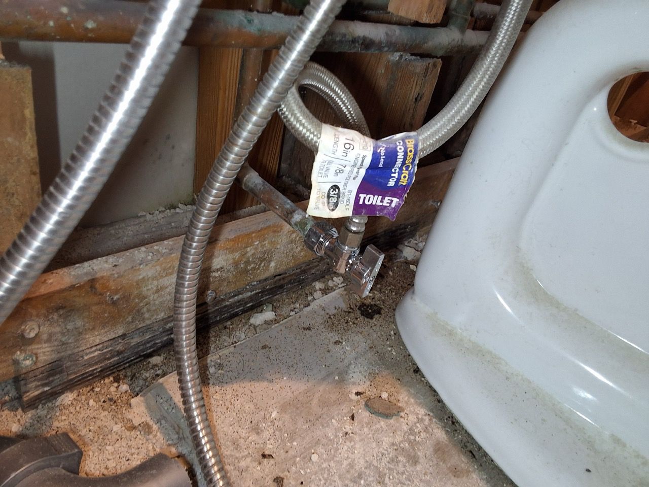 Silver braided hoses with a purple tag beside a white appliance in a rusty under-sink area