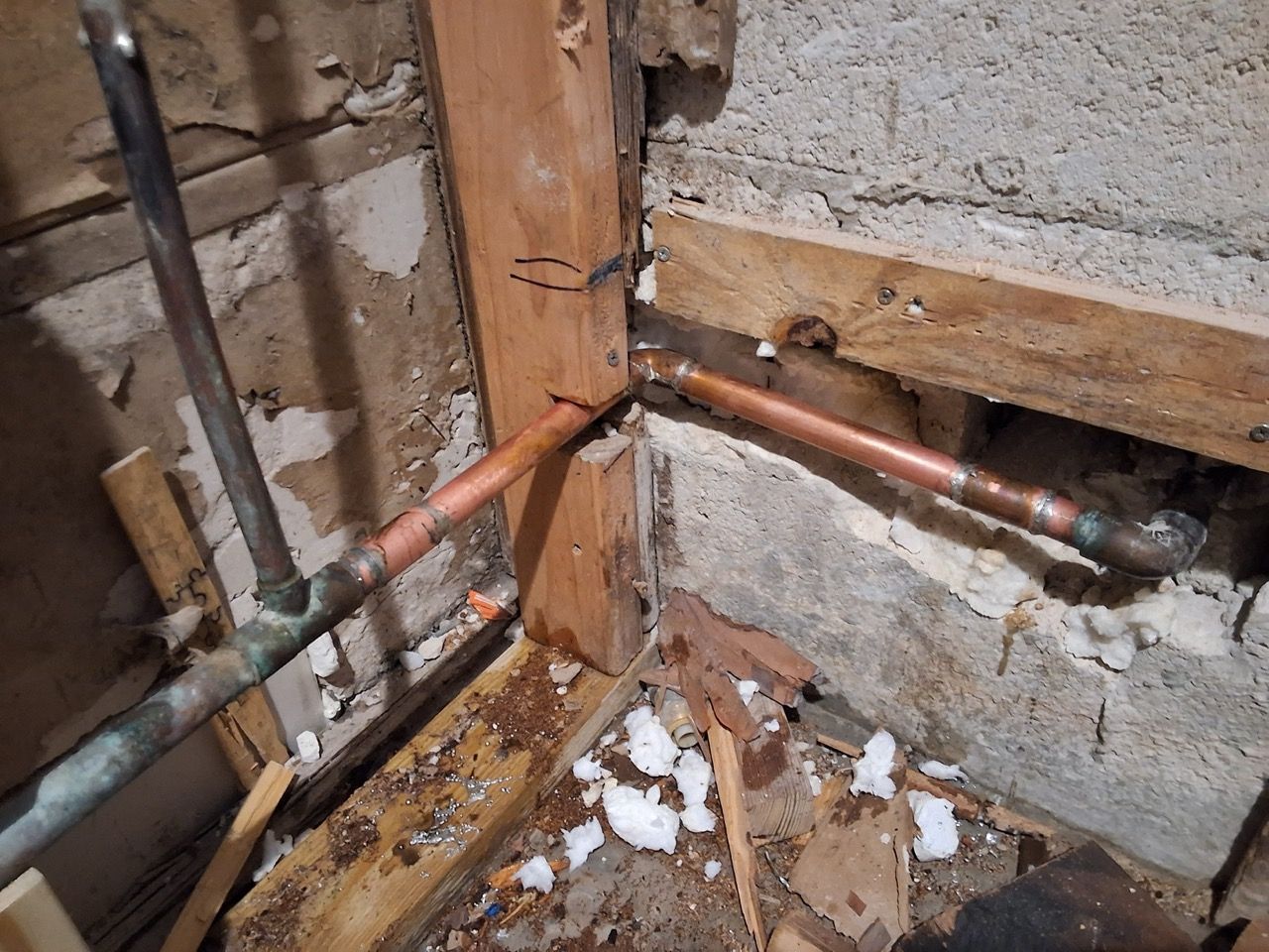 Exposed copper pipes and damaged wall framing in a water-damaged basement corner with debris on the floor