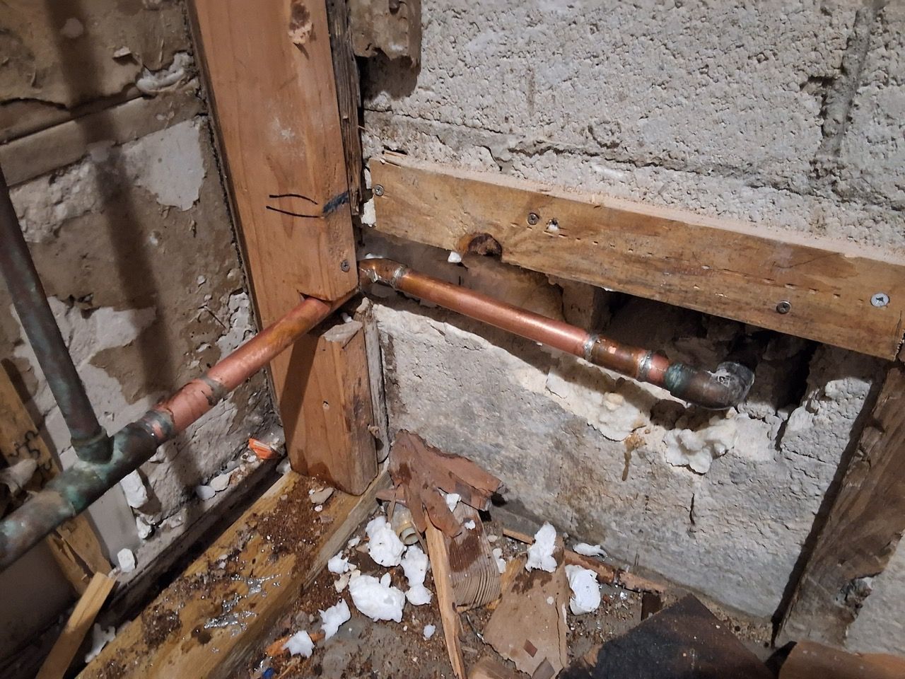 Damaged wall corner with exposed copper pipes and debris on the floor