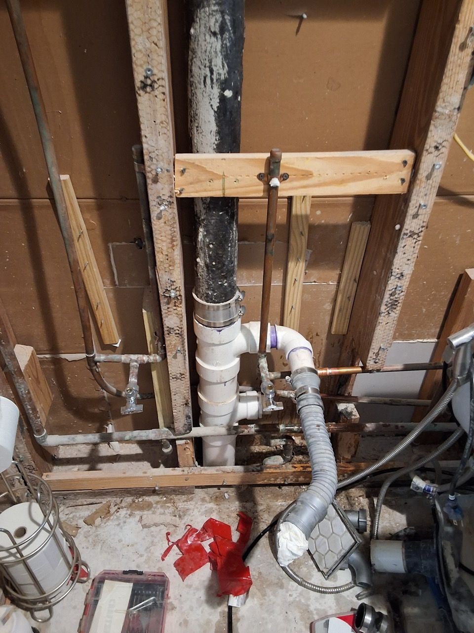Exposed plumbing and drain pipes inside a wall frame, with a red stain on the floor below.