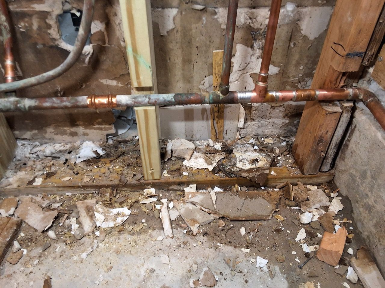 Damaged wall with exposed pipes, torn insulation, and debris on a rough concrete floor.