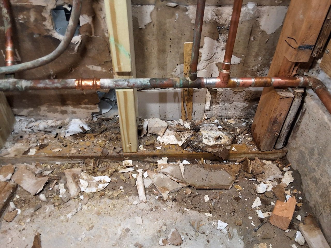 Damaged wall with exposed pipes, broken plaster, and debris on the floor in a construction area