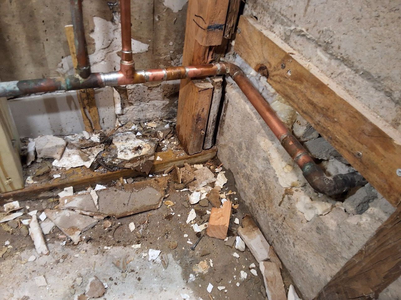Damaged basement corner with exposed pipes, broken plaster, debris, and water stains on walls and floor