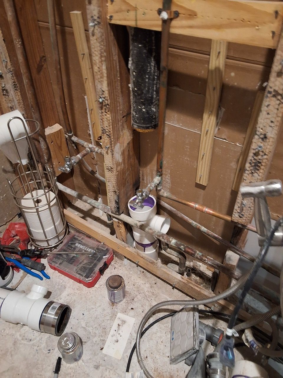 Exposed plumbing and wooden wall studs in a renovation area with scattered tools and pipes on the floor