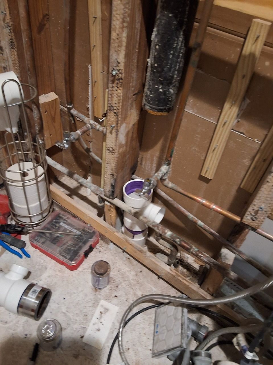 Plumbing under a sink with exposed pipes, a white water filter, and tools on the floor.