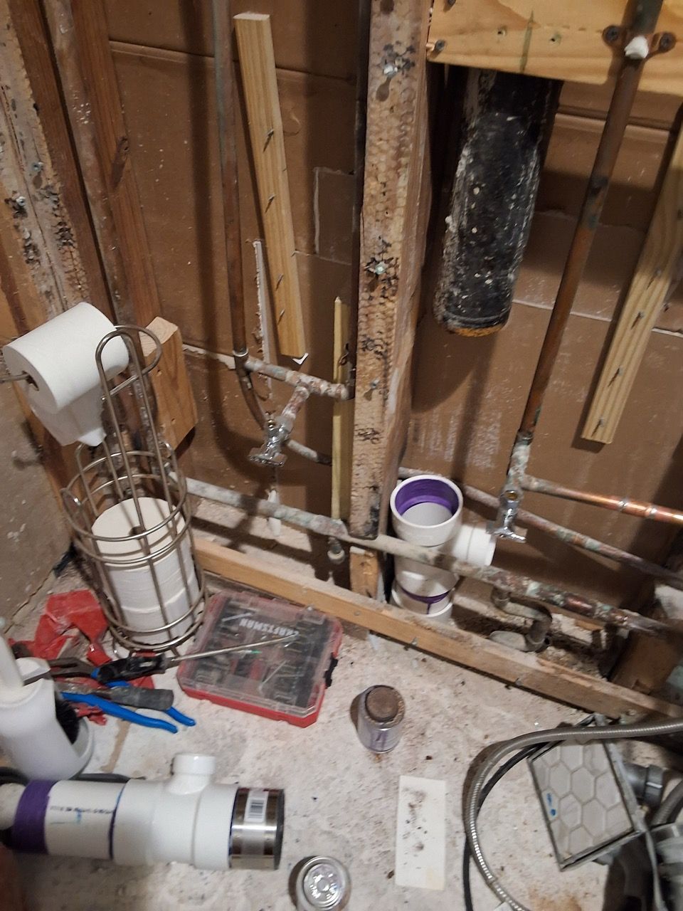 Exposed plumbing and pipes inside a wall, with tools and a white bucket on a construction floor.